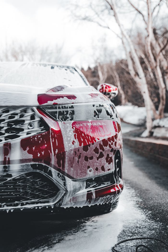 Close-up view of a red car covered in foam during a car wash. The image showcases soap suds on the grille and headlight